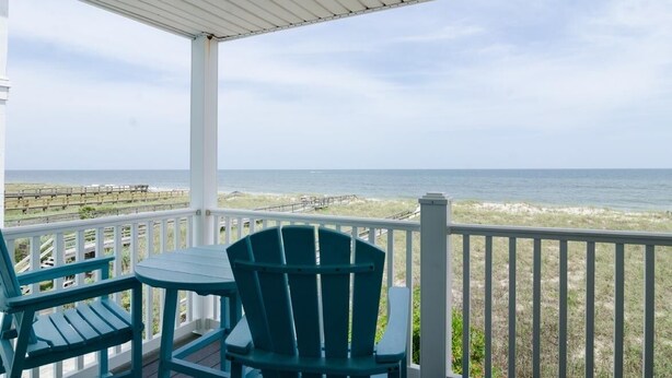 Een overdekt balkon met uitzicht op de oceaan, met twee groene Adirondack-stoelen en een tafeltje.
