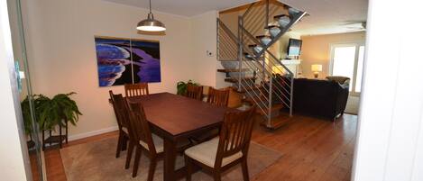 A dining area with a wooden table and chairs, a staircase with a metal railing, and a painting on the wall.