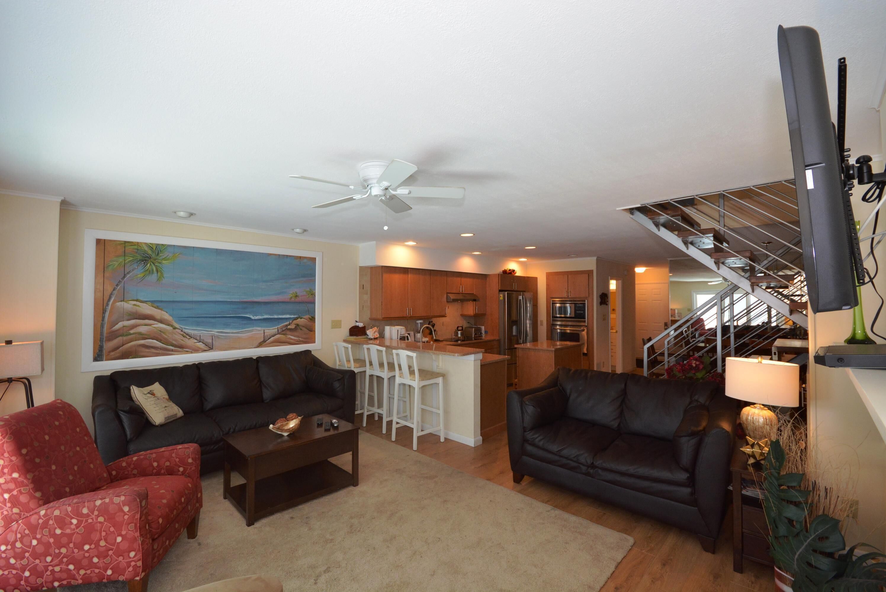 A living room with a sofa, armchair, coffee table, and a painting of a beach scene. There is a kitchen area with a bar and a staircase leading to an upper level.