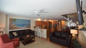 A living room with a sofa, armchair, coffee table, and a painting of a beach scene. There is a kitchen area with a bar and a staircase leading to an upper level. - Unique, stylish 4 bedroom bayside townhouse with free WiFi, pier boat docking, and a breathtaking view of the ocean located midtown on the bay water and just a few blocks from the beach! (Ocean City)