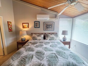 Une chambre à coucher comprenant un lit, deux tables de chevet, un ventilateur de plafond, un appareil de climatisation et une fenêtre avec des stores.