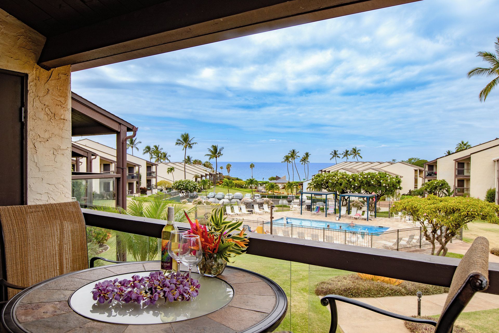 A balcony with a table set for two, overlooking a pool area with lounge chairs and a view of the ocean.