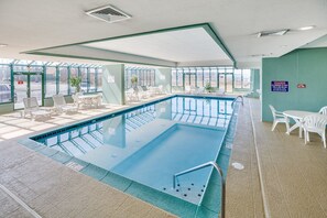 Una piscina interior con paredes de vidrio transparente, sillones blancos para relajarse y techo verde.