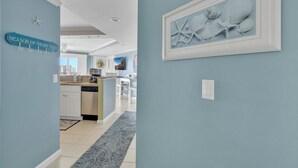 Un couloir aux murs bleu clair, un coin cuisine avec des meubles blancs, un tapis gris et une œuvre d’art représentant un coquillage, encadrée.