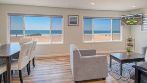 Ein Zimmer mit großen Fenstern mit Blick auf einen Strand, einem Essbereich mit Stühlen, einem Sofa und einem Fernseher an der Wand.