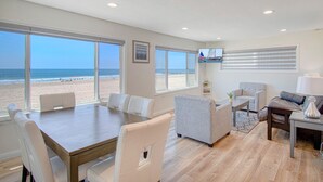 Ein geräumiges Wohnzimmer mit großem Fenster und Blick auf einen Strand, einem Essbereich mit Holztisch sowie einer Sitzecke mit Sofa und Sesseln.