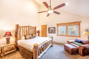 A bedroom with a rustic wooden bed frame, a ceiling fan, a window with a view, and a leather armchair.