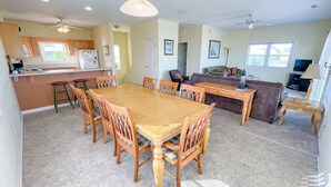 Een ruime woonkamer met een eettafel en stoelen, een keuken met een koelkast, en een zithoek met een bank en stoelen.