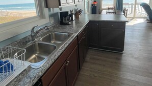 A kitchen with granite countertops, stainless steel sinks, and wooden cabinets. There is a large window providing a view of the beach. - Island Delights II – Sun, Sand & Simple Fun (Surf City)