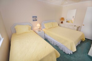 A room with two beds, each with a white headboard and a yellow bedspread. There is a nightstand with a lamp and a small table with a lamp on it. A mirror is on the wall above the bed.