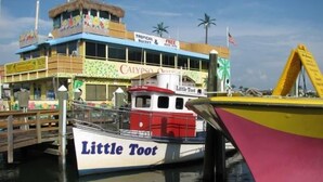 Un bote colorido llamado 'Little Toot' atracado en un muelle con un edificio al fondo identificado como 'Calypso Marina' y 'Tropical Buffet & Free Beer'.