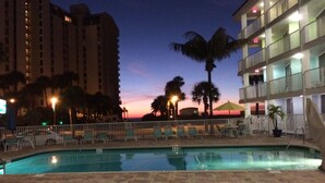 Zona de piscina con sillones de descanso y una valla, rodeada de palmeras y edificios.