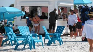 Un quiosco de comida en la playa con sillas y sombrillas azules, rodeado de gente.