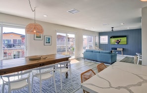 A modern living room with a blue sofa, a wooden dining table with white chairs, a television mounted on the wall, and a balcony with a railing.