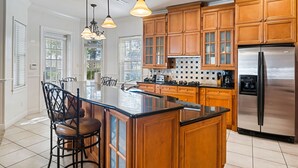 A kitchen with wooden cabinets, a stainless steel refrigerator, a large island with bar stools, pendant lights, and a window with blinds. - Homestead Holly: Game Loft, Private Pool, Water Park, Near Disney (Kissimmee)