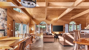 A spacious wooden interior with a long dining table, sofas, and a TV.