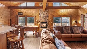A log cabin interior with a stone fireplace, wooden furniture, and large windows.