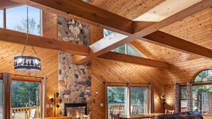 A wooden cabin interior with a stone fireplace, large windows, and a high ceiling.