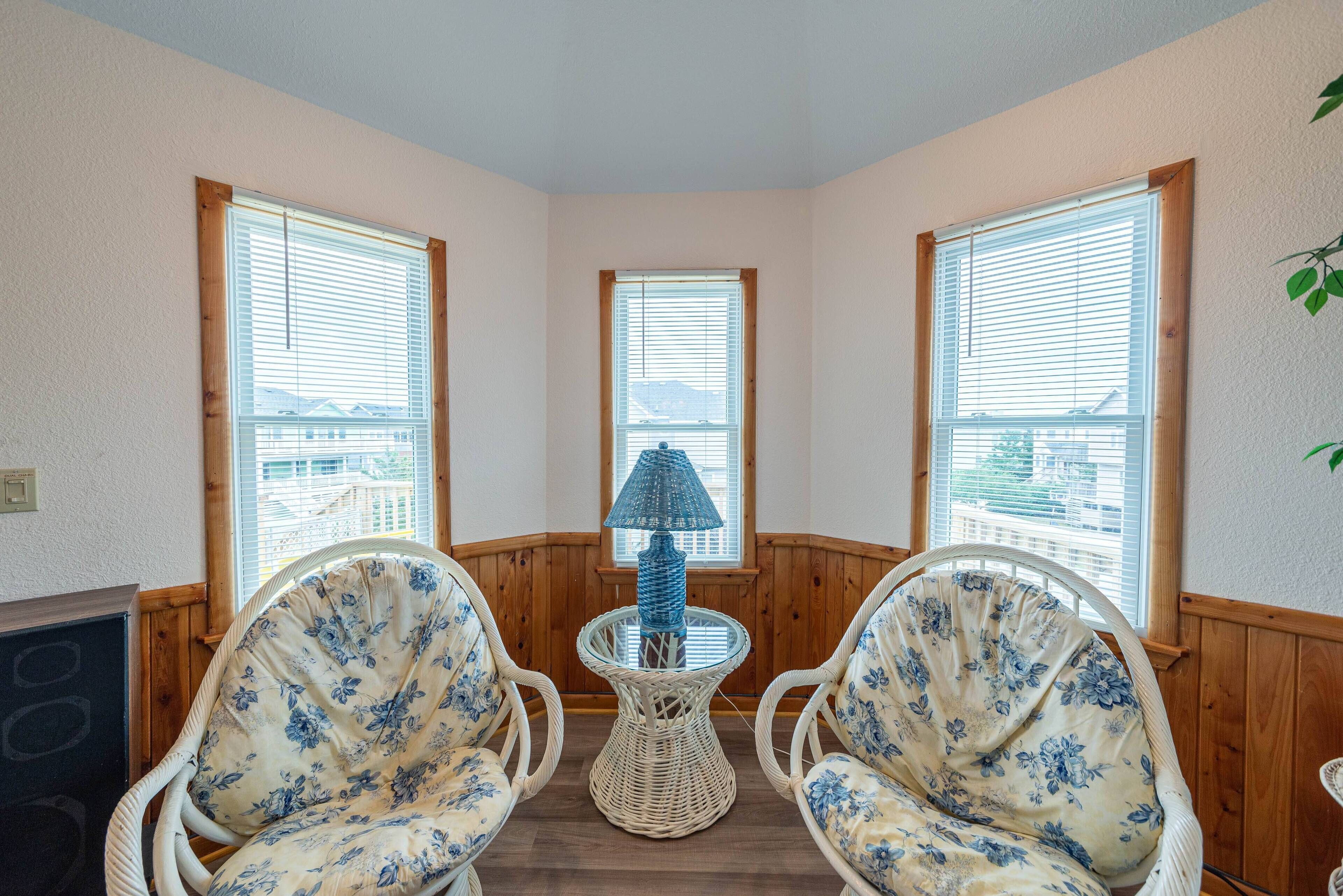 A corner room with two wicker chairs, a small round table with a lamp, and large windows with blinds.