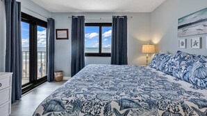 A bedroom with a large bed, a window with curtains, a bedside lamp, and a view of the beach.