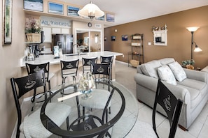 A modern living room with a round glass table, black chairs, a grey sofa, and a kitchen area with a fridge and coffee maker.