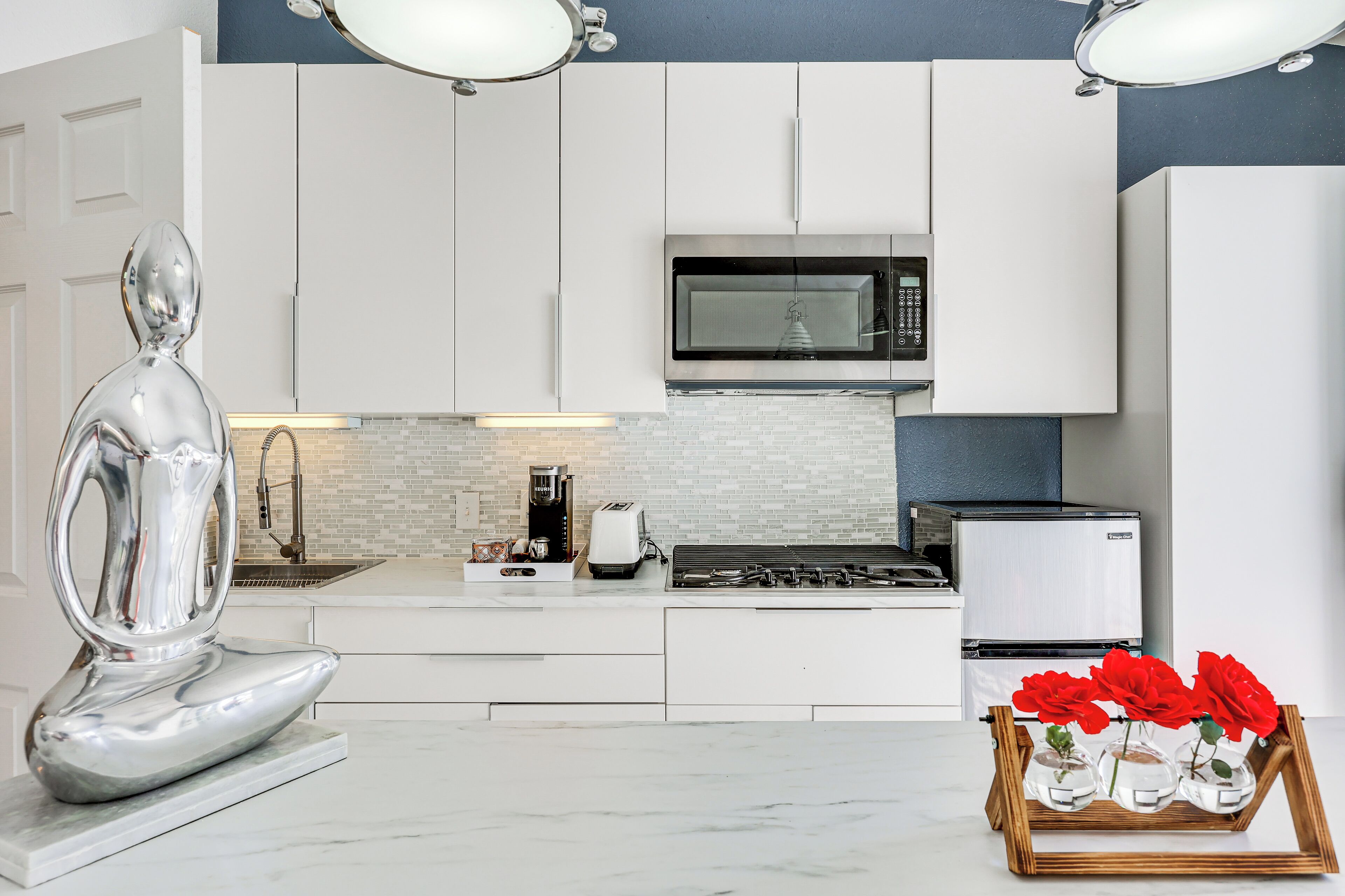 A modern kitchen with white cabinets, a microwave, a stove, and a coffee maker. There is a decorative silver sculpture and a tray with red roses.