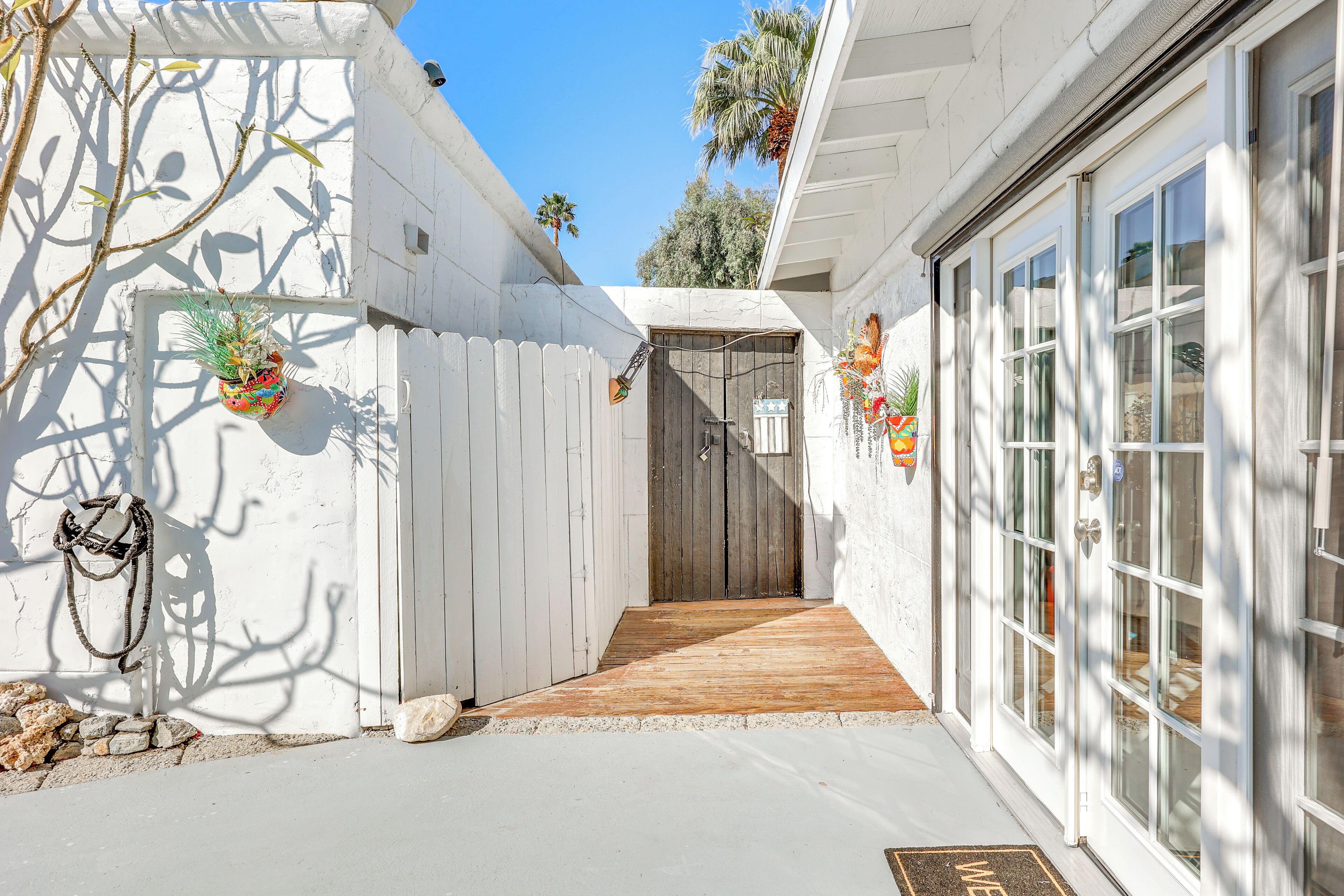 A narrow pathway leading to a wooden door, flanked by white walls and a white fence.