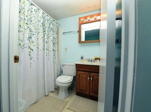 A bathroom with a toilet, a wooden cabinet, a mirror with lights, and a shower curtain with a leaf pattern. - Spacious, comfy 2 bedroom condo with free WiFi and a wide balcony located midtown on the ocean block just a short walk to the beach! (Ocean City)