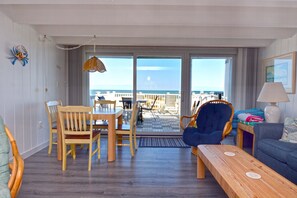 Quarto com mesa e cadeiras de madeira, uma poltrona azul, um sofá e um abajur. Vê-se a praia através das portas de vidro.