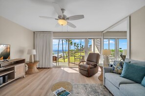 A modern living room with a ceiling fan, a sofa, a TV, and a view of the ocean through large windows. - Kapaa Shore Resort C227, Oceanfront, End-Unit, AC, Washer/Dryer, Bike Path (Kapaa)