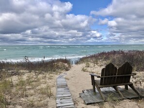 A wooden bench facing a beach with a wooden boardwalk leading to the water. - Unique round home Lake Michigan waterfront. Sunset views, fire pit, dog ok. (Northport)