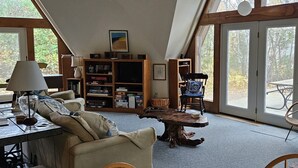 Uma sala de estar acolhedora com um sofá, uma mesa de centro e uma estante de livros. Há uma janela grande que oferece luz natural e vista para o exterior.