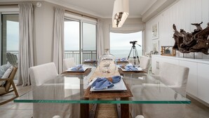 Un comedor moderno con una mesa de vidrio, sillas blancas tapizadas y vistas al océano a través de amplios ventanales.