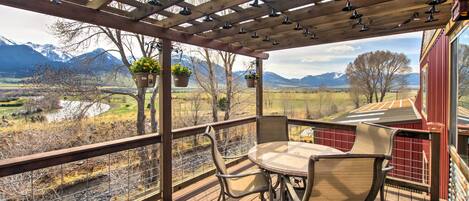 A wooden deck with a table and chairs, overlooking a scenic landscape with mountains and a river.