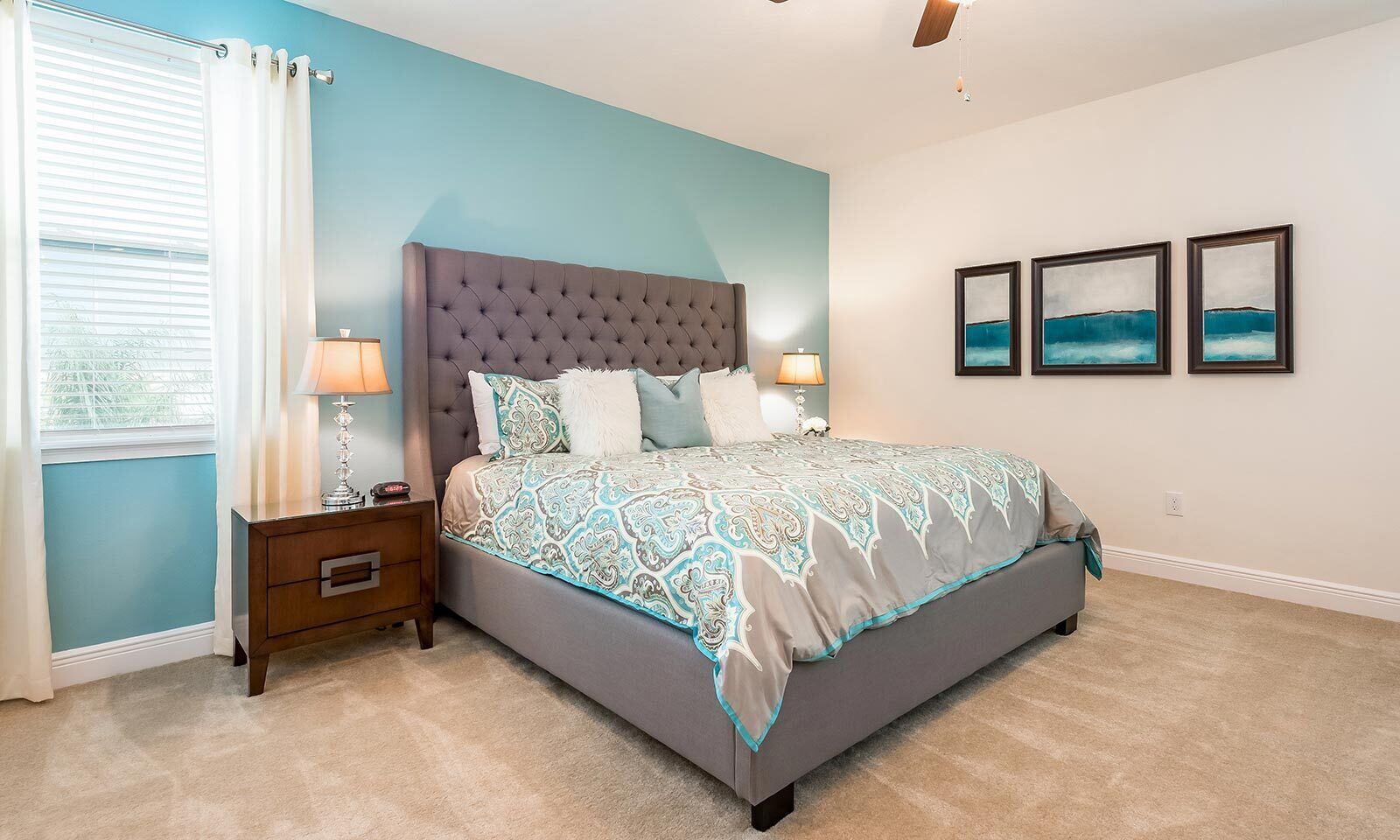 A bedroom with a tufted headboard, a patterned bedspread, a nightstand with a lamp, and a window with curtains.