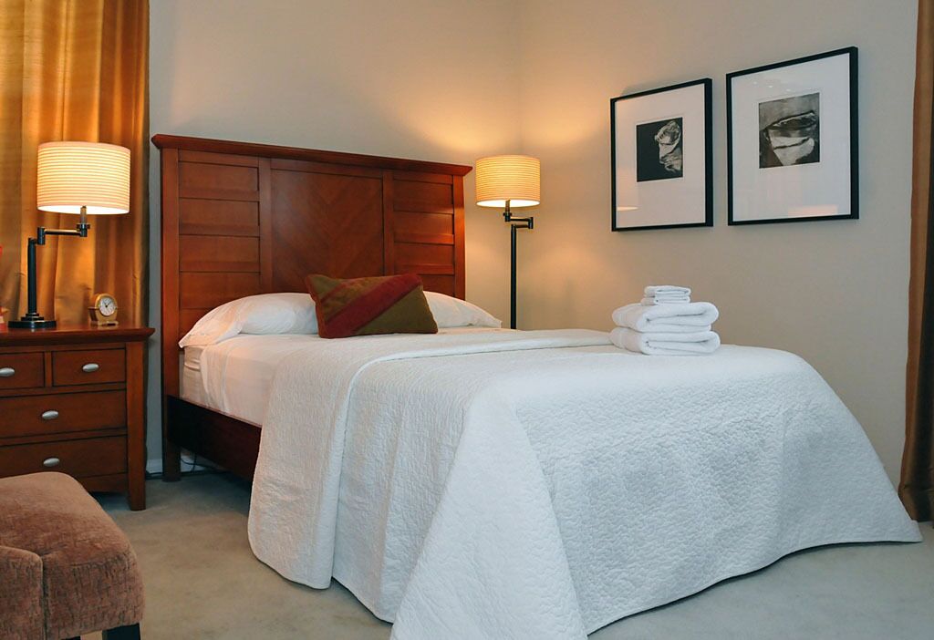 Een hotelkamer met een houten hoofdeinde, twee bedlampen, een net opgemaakt bed met witte lakens, een bruine bank en twee ingelijste foto’s aan de muur.