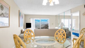 Un área de comedor con una mesa de vidrio y sillas de madera, un sofá, un televisor y un balcón con vista a la ciudad.