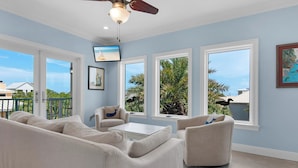 A spacious living room with a ceiling fan, large windows with views of palm trees, and a beige sectional sofa. - 0.2 Mi to Beach, Tiki Bar, Huge Pool, Close to Silver Sands Shopping (Miramar Beach)