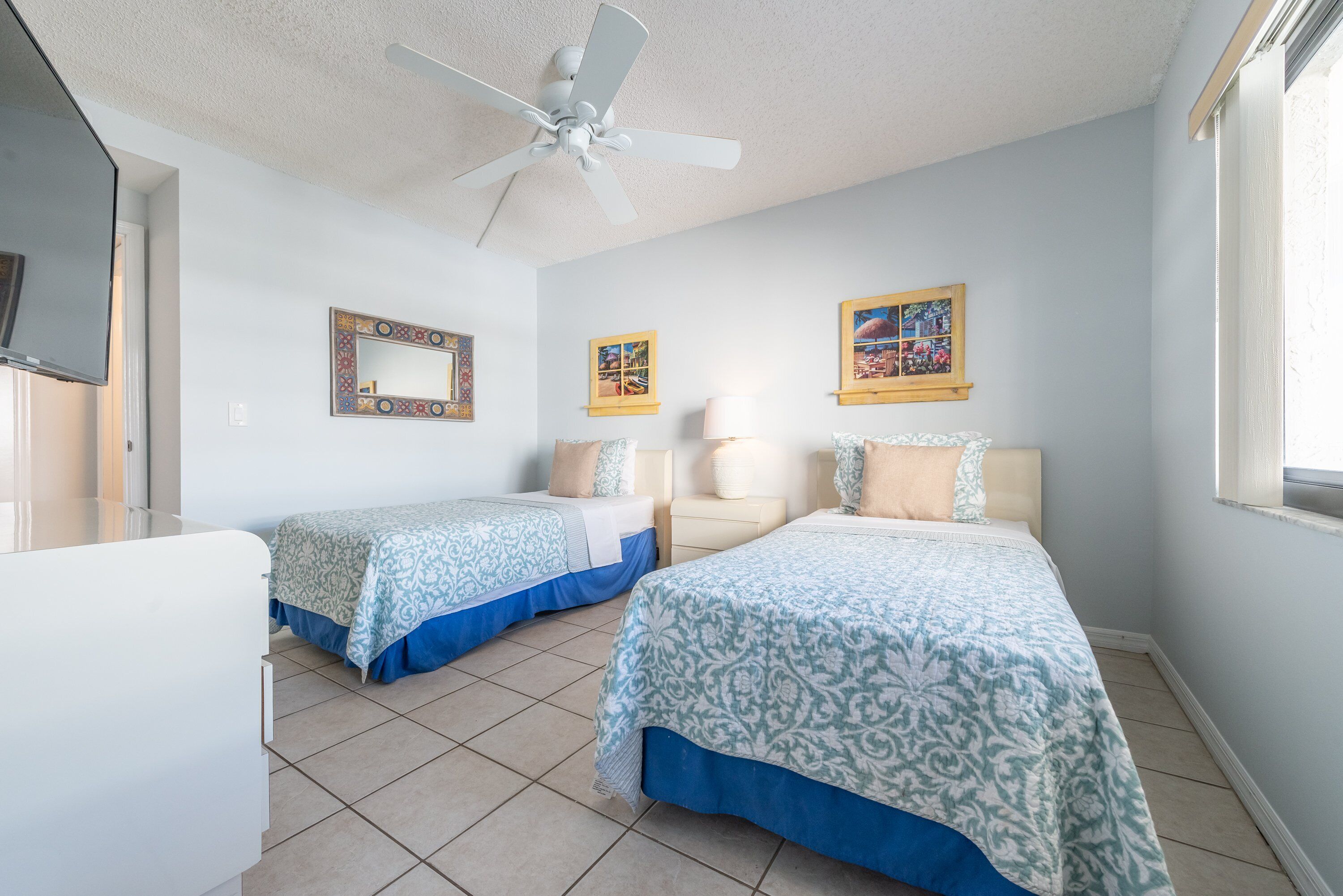 A hotel room with two beds, a ceiling fan, a TV, and artwork on the walls.