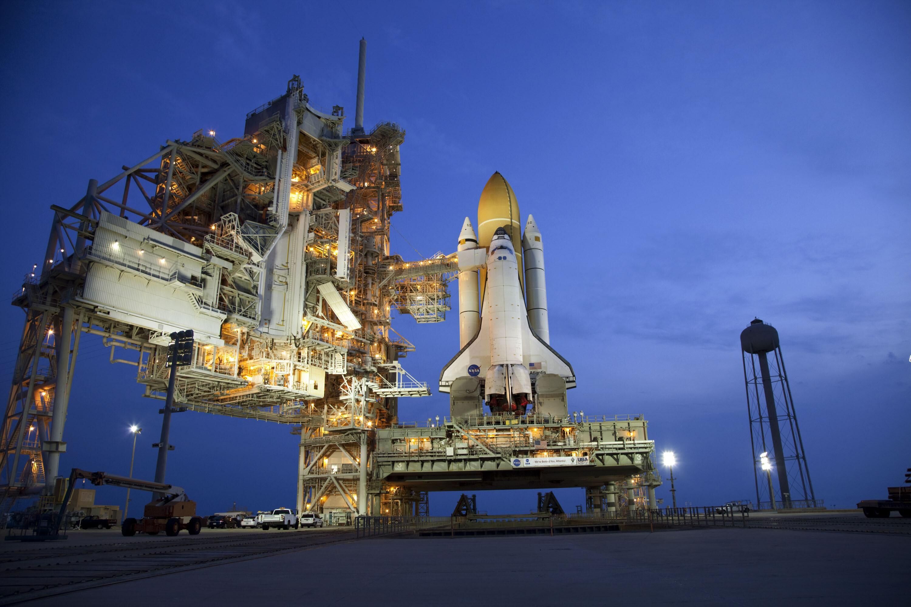 Un transbordador espacial sobre una plataforma de lanzamiento, con una gran estructura industrial y una torre de agua al fondo.