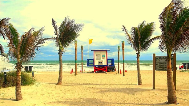 Een strand met een redderpost, palmbomen en een bord.