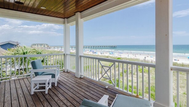 A wooden deck with white railings overlooking a beach and pier.