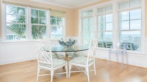 A dining area with a glass table surrounded by white chairs, a ceiling fan, and large windows with views of palm trees.