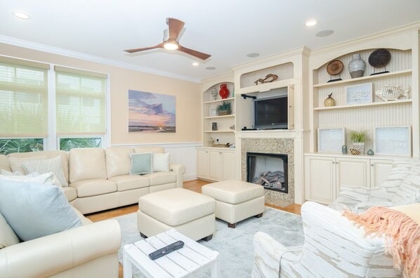 A spacious living room with a beige sofa, ottomans, a fireplace, and a ceiling fan. - Grand 5BR oceanfront home w private beach access! (Wrightsville Beach)