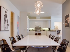 A modern dining room with a large white table and chairs, a kitchen in the background, and artwork on the walls.