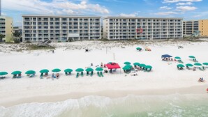 A beach with multiple green umbrellas and lounge chairs, a red tent, and a large building in the background. - GD 307: Seaglass Cove - Step Out, Chill Out... Beachfront Living at Its Best! (Fort Walton Beach)