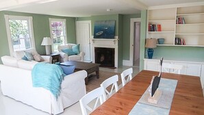 A spacious living room with a fireplace, a dining area with a long wooden table, and a bookshelf filled with books. - Charming house located in the heart of downtown Chatham! (Chatham)