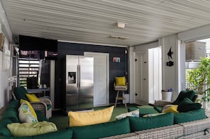 Un espace de vie extérieur abrité, comprenant un canapé, des fauteuils, un réfrigérateur et une porte donnant sur une autre pièce.