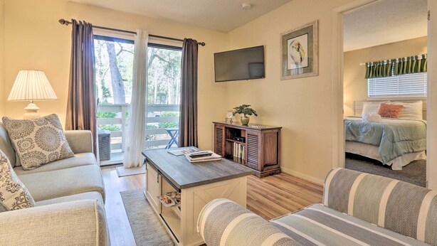A cozy living room with a sofa, armchair, and a coffee table. There is a TV mounted on the wall and a window with curtains. - Walk to Beach: Resort-Style Hilton Head Condo! (Hilton Head Island)