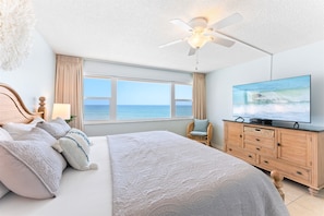 A bedroom with a bed, a television, a ceiling fan, a window with a view of the ocean, and a wooden dresser.